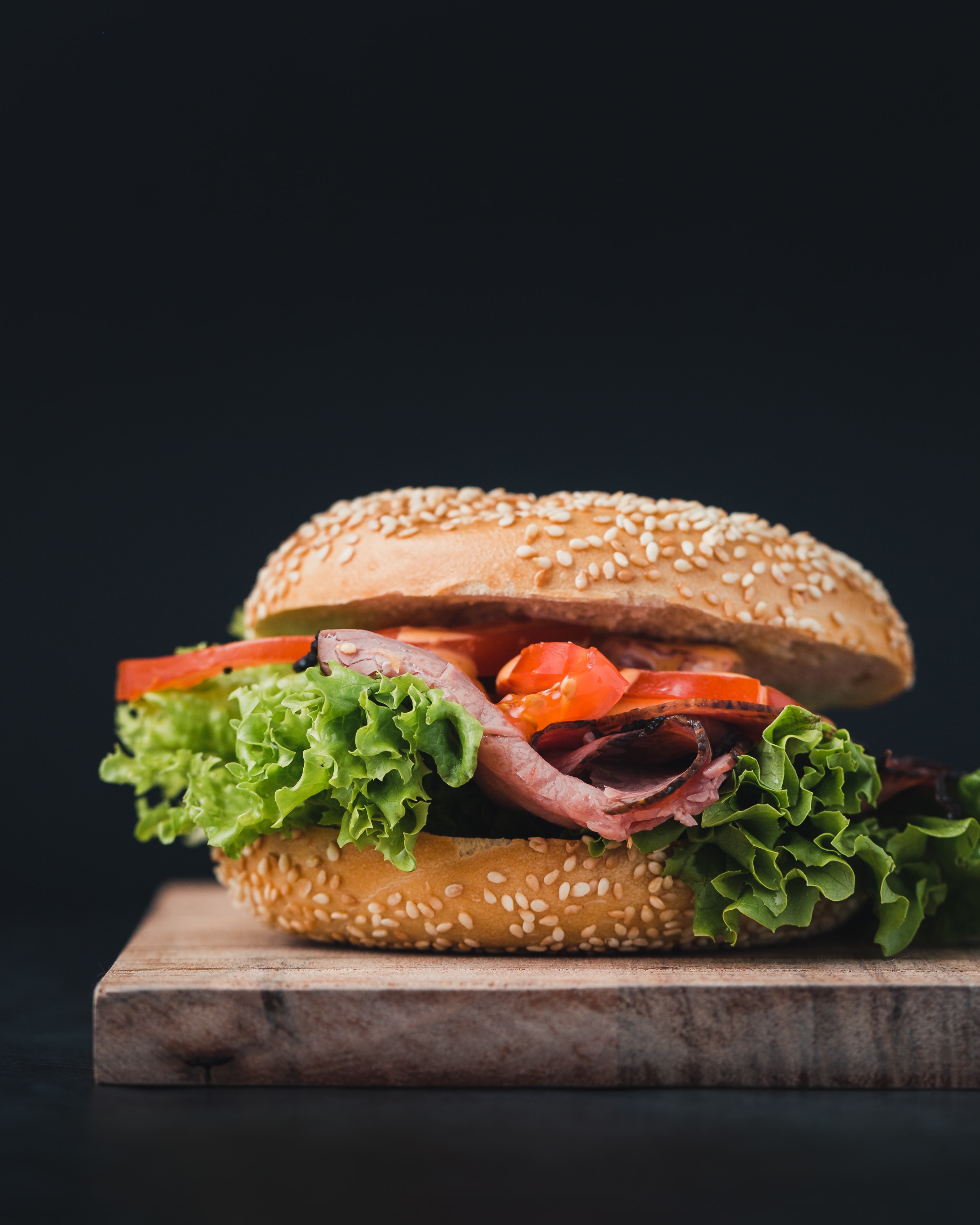 Against a moody black background, there is a sloppy burger on a sesame seed bun, comprised of some deli meat, lettuce, and tomato. The entire burger is placed on a wood cutting board and the photo has a very shallow depth of field, so the back of the cutting board is blurry.