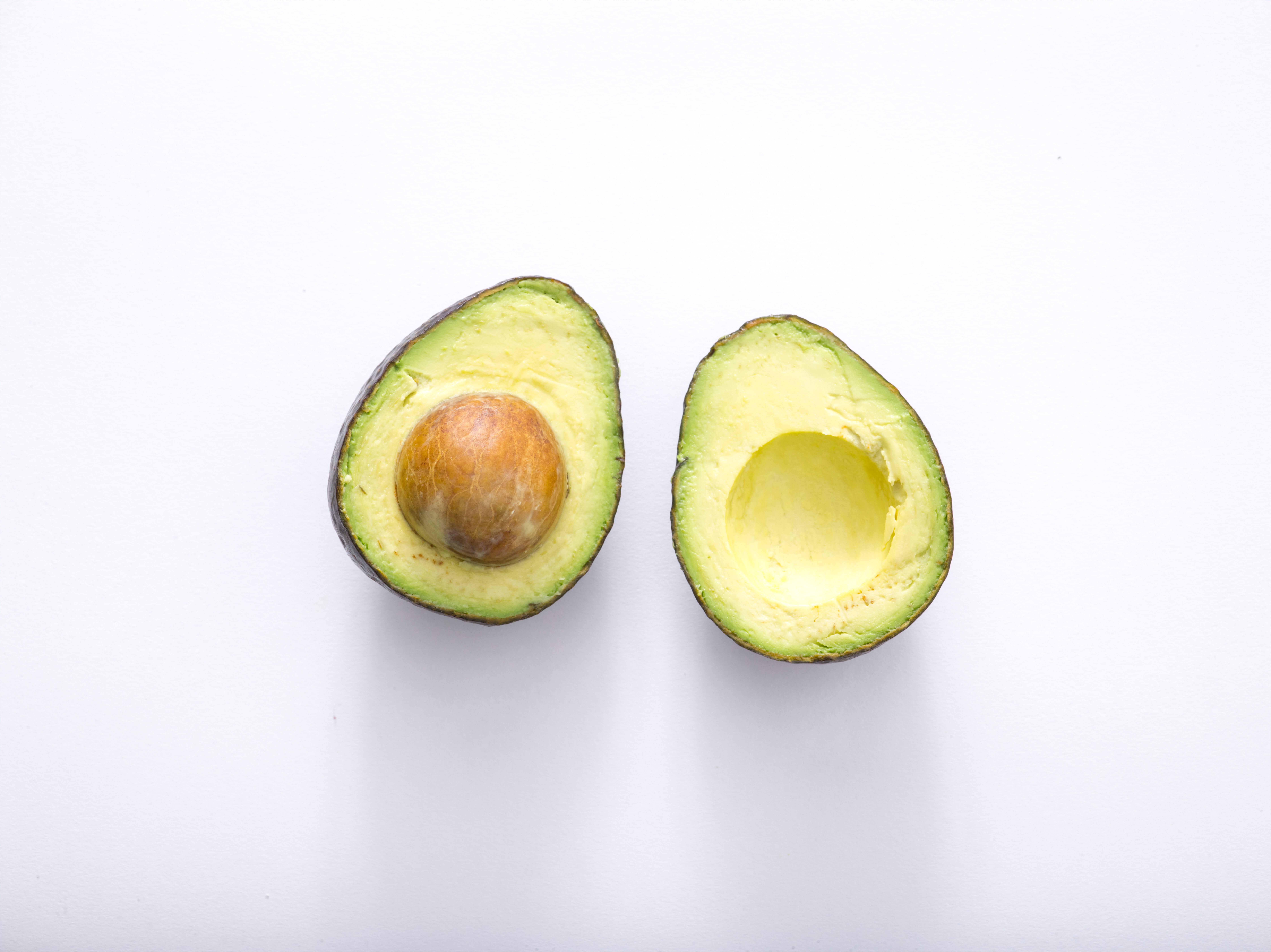 Two halves of a small Haas avocado appear side by side, skin side down, on a white background. The left avocado still has the pit.